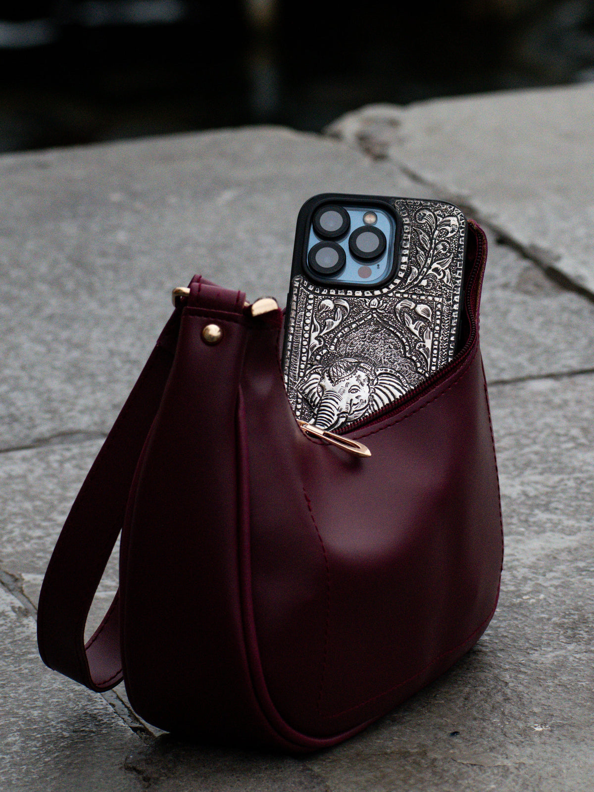 A burgundy leather shoulder bag with gold-tone hardware sits on a stone dock with a blurred waterway and boat in the background. A smartphone with a silver patterned case is tucked into the bag's front pocket.